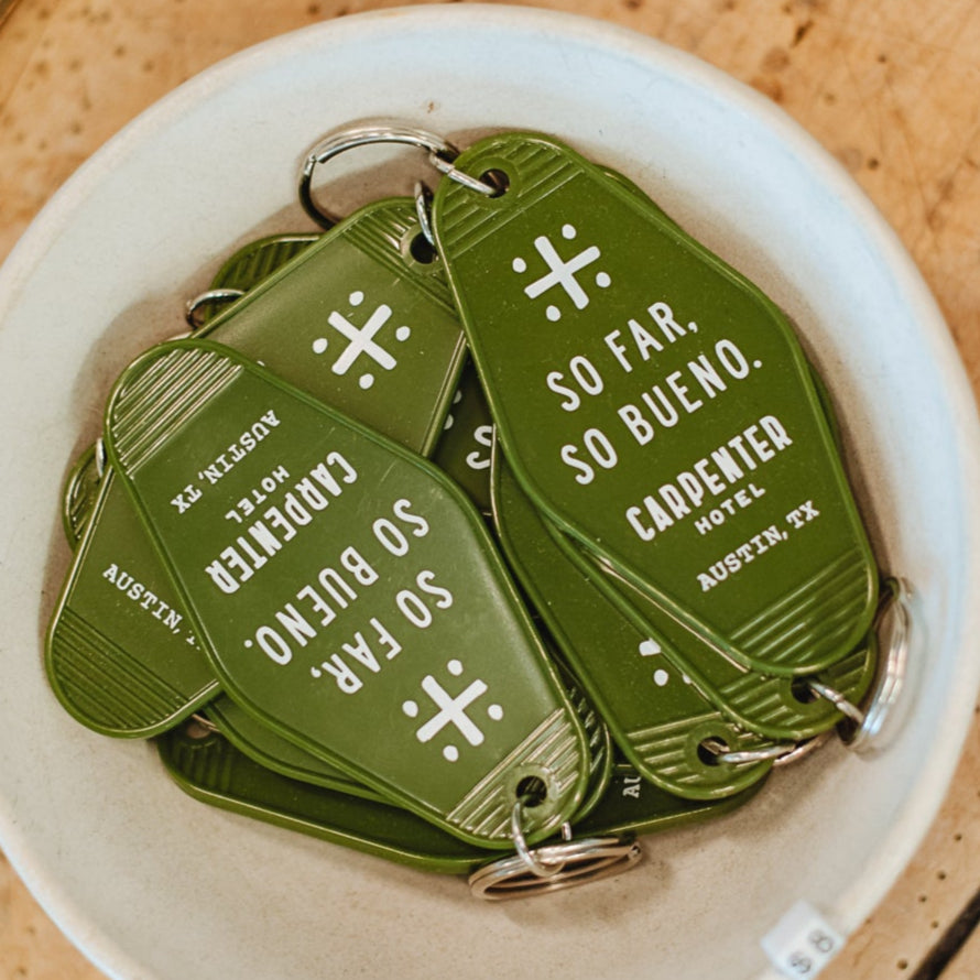 Green vintage-style keychains with white text that reads 'SO FAR, SO BUENO CARPENTER HOTEL AUSTIN, TX'. in a white bowl sitting on top of a wooden table