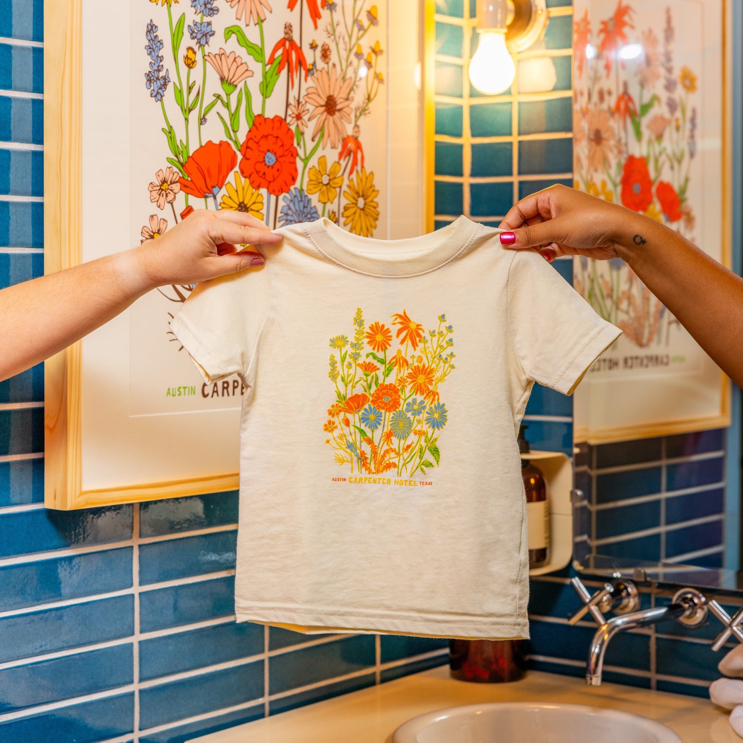 A beige kids' t-shirt with a colorful floral graphic design and text that says ' CARPENTER HOTEL AUSTIN TEXAS' on the chest being held up by two hands with matching poster hanging in background against blue tile.