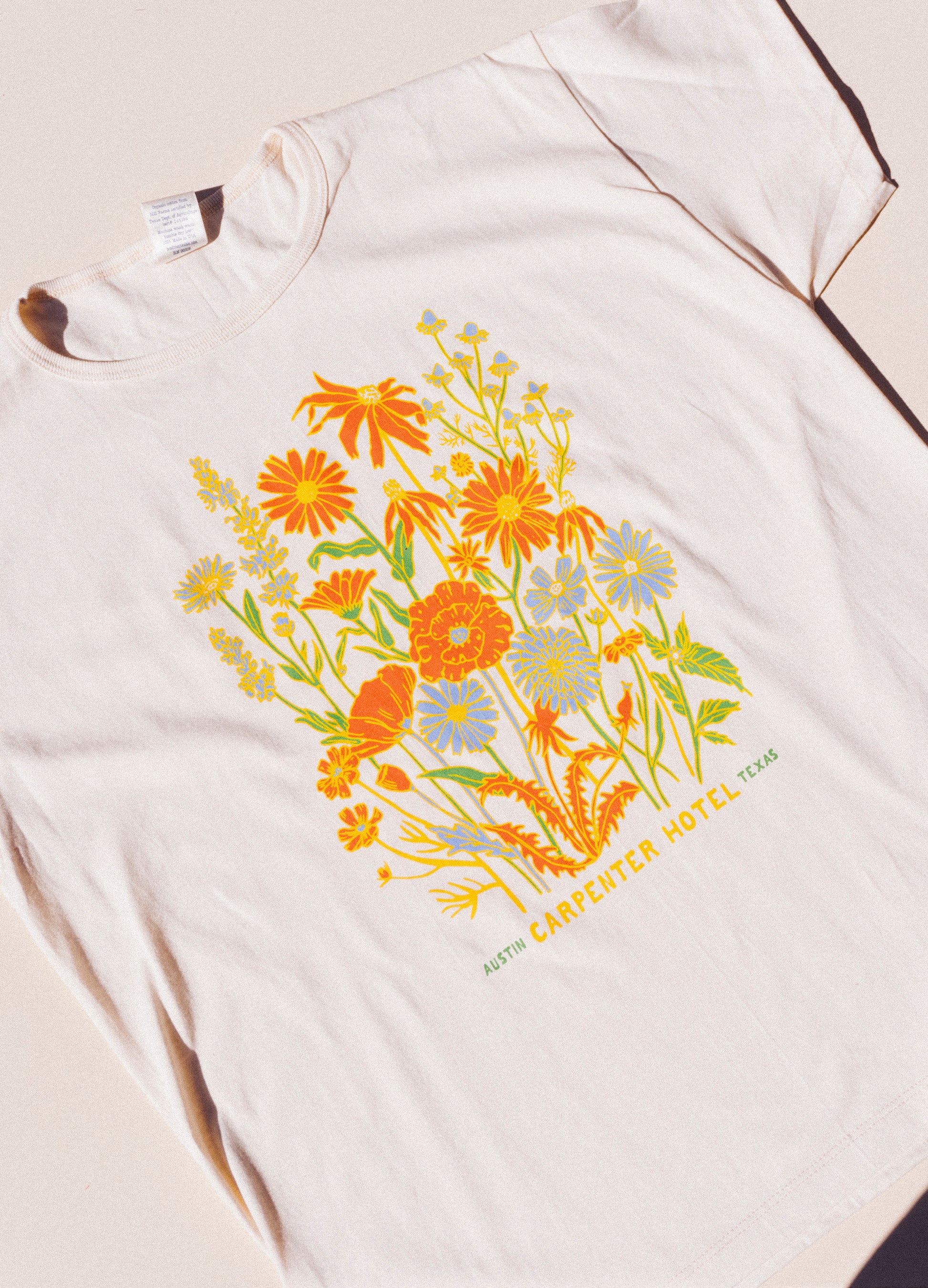 Photograph of beige cotton T-shirt with colorful floral print with text below it that reads 'CARPENTER HOTEL AUSTIN TEXAS' laying on top of beige background lit by natural sunlight